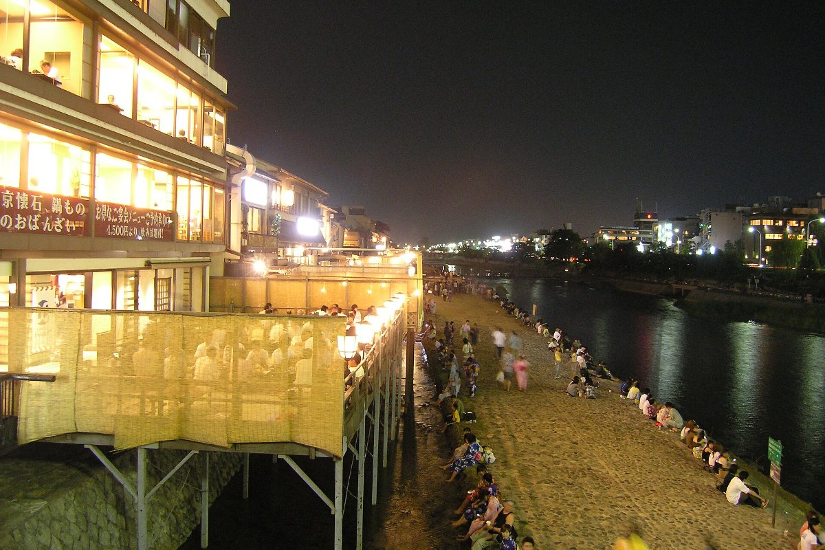 ระเบียงไม้ของร้านอาหารเรียงริมแม่น้ำคาโมะ เปิดไฟยามเย็น มีคนนั่งกินข้าวบนระเบียงย่านพนโตะโจ เกียวโต