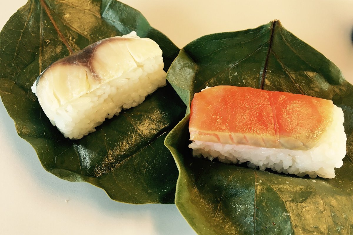 ซูชิกดชิ้นเล็กห่อด้วยใบพลับสีเขียวเรียงในกล่องไม้ แกะใบออกให้เห็นปลาซาบะบนข้าว นารา