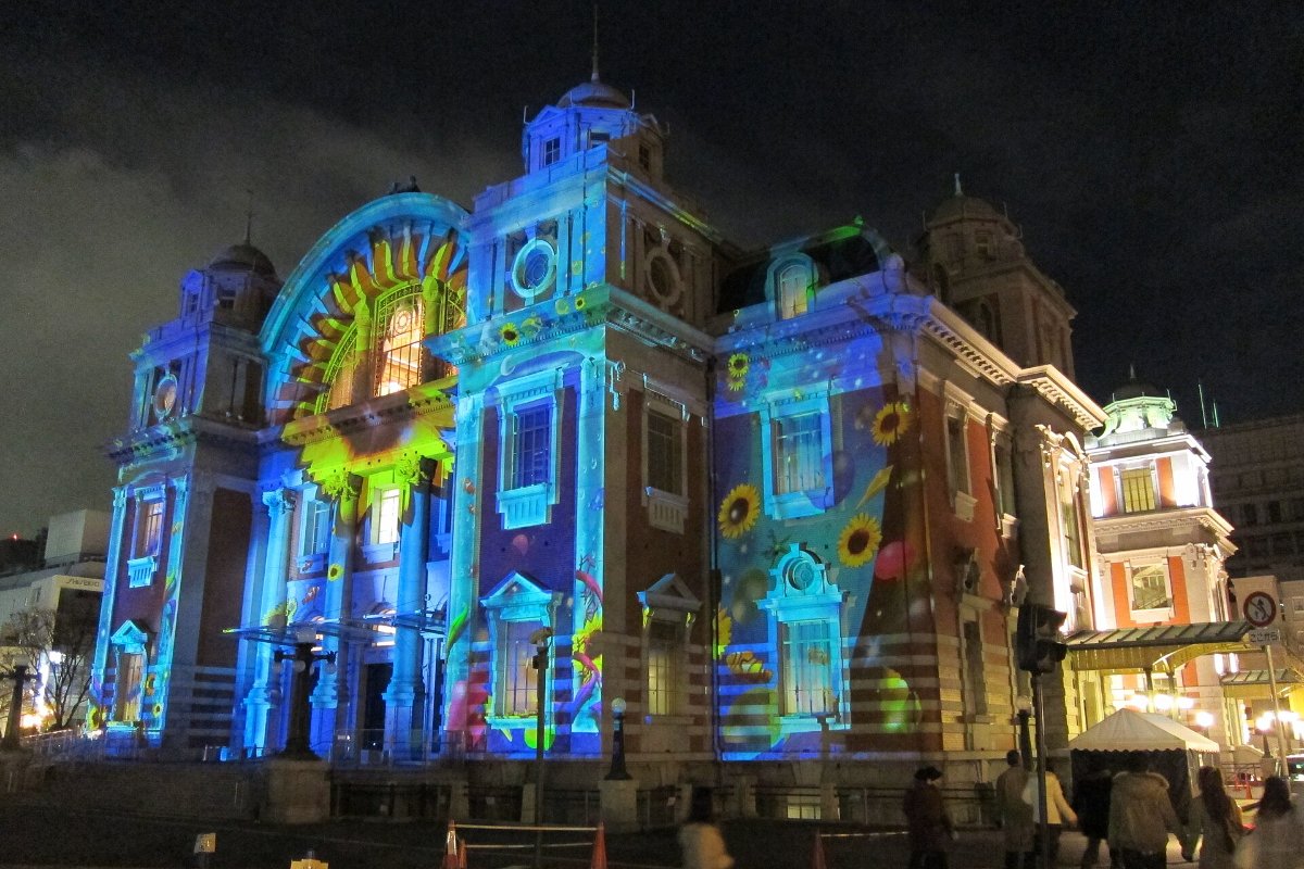 ภาพฉายแสงสีบนผนังด้านหน้าอาคารอิฐแดงหอประชุมกลางเมืองโอซาก้าตอนกลางคืน มีผู้ชมยืนดูบนเกาะนากาโนชิมะ